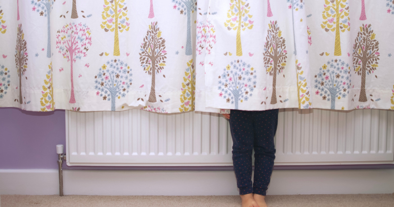 A girl hiding behind a long curtain adjacent to a wall-mounted electric radiator.