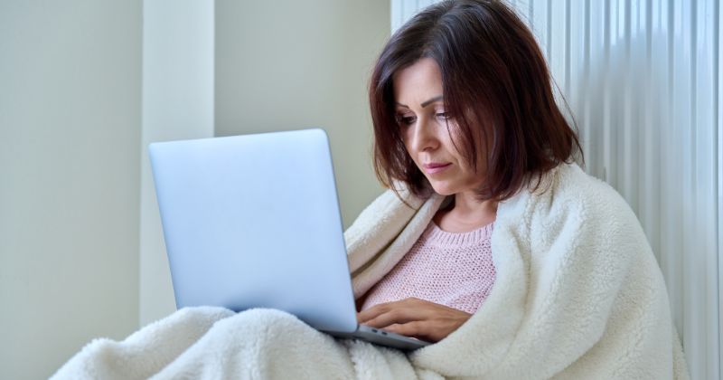 A woman, working on her laptop, is keeping warm next to a wall-mounted radiator.