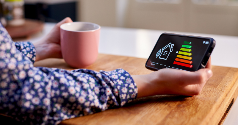 Close-up of a woman holding a smart energy meter in the kitchen, measuring energy efficiency while enjoying a cup of coffee.