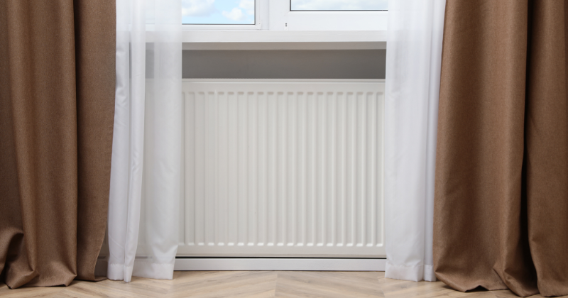 White wall-mounted electric radiator under window with white and brown curtains.